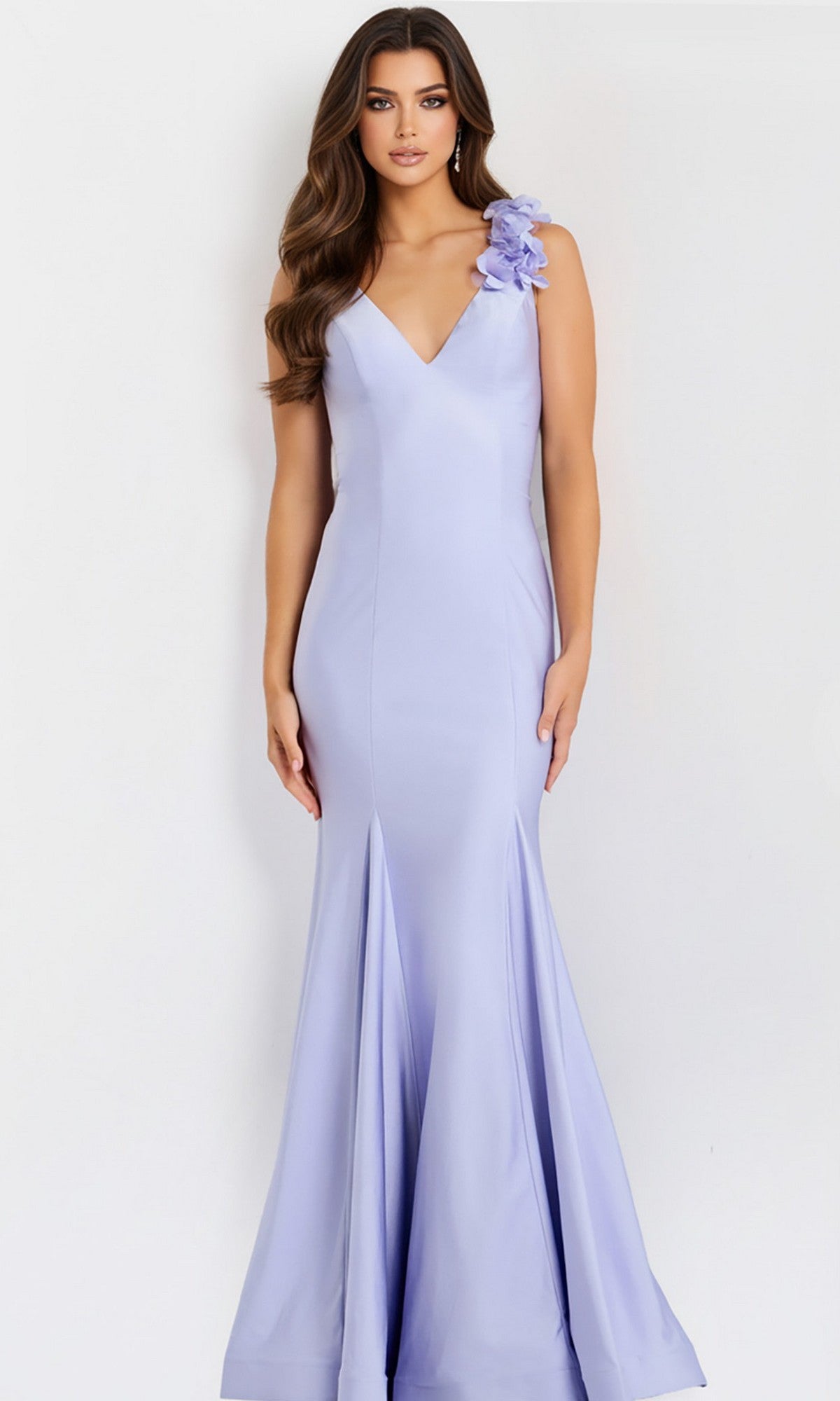 A woman poses against a plain light background wearing the Jovani 48120 long prom dress, featuring a fitted, floor-length silhouette with a V-neckline and 3D floral appliques on one shoulder.